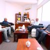 Five men sit in a lounge-style office, chatting around a wooden coffee table with cups and snacks nearby.