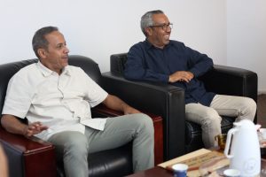 Two men sit on black leather couches in a bright lounge, smiling and talking.