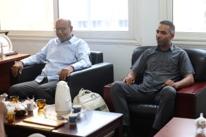Two men in casual shirts sit on black leather couches in a bright office lounge, with a coffee table of cups and snacks in front of them.