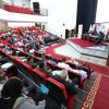 Audience-filled conference hall with red seats watching a panel on stage with banners and flags on the right side(vivid, wide-angle view).