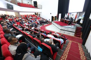 Audience-filled conference hall with red seats watching a panel on stage with banners and flags on the right side(vivid, wide-angle view).