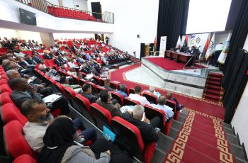 Audience-filled conference hall with red seats watching a panel on stage with banners and flags on the right side(vivid, wide-angle view).