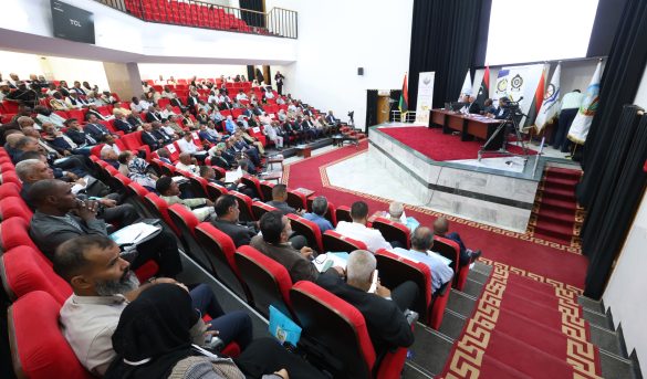 Audience-filled conference hall with red seats watching a panel on stage with banners and flags on the right side(vivid, wide-angle view).