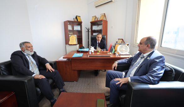 Three men in business suits sit around a wooden desk in a bright office, having a formal meeting with awards and flags on the desk.