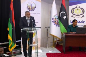 Man in a suit speaking at a glass podium during a formal event, with Libyan flag and banners in the background.