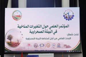 Large conference slide on a screen featuring Arabic text and several organizational logos at the top corners, with decorative circular images on the left and bottom edges.