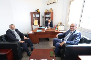 Three men in suits sit around a wooden desk in a bright office with award plaques on display shelves behind them.