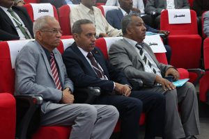 Three men in suits sit in a row of red theater seats at an event, wearing lanyards and looking ahead.