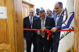 Officials in suits cut a red ribbon during a formal ribbon-cutting ceremony beside a wooden-paneled hallway and a flag in the background.