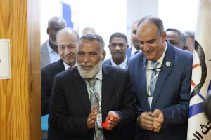 Men in suits cutting a red ribbon at a ceremonial event, with others smiling in the background and flags nearby.
