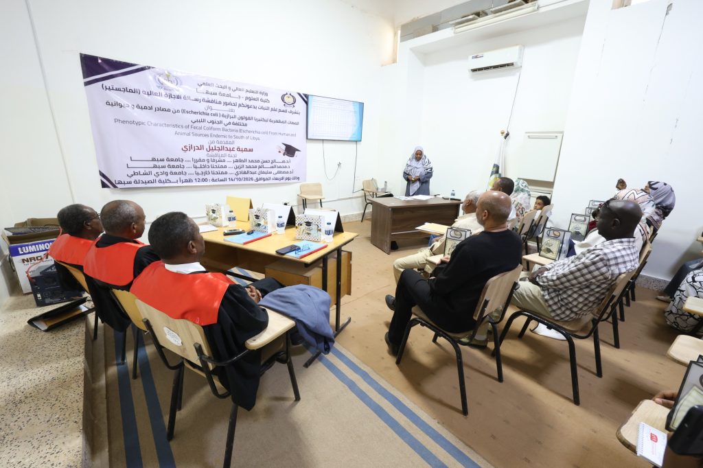 Audience seated in a classroom watching a presenter at a desk, with a large Arabic banner on the wall behind them.