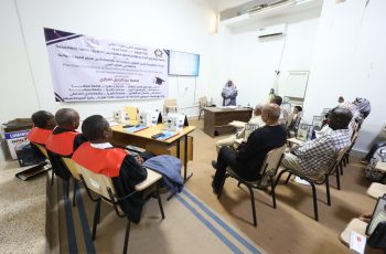 Audience seated in a classroom watching a presenter at a desk, with a large Arabic banner on the wall behind them.