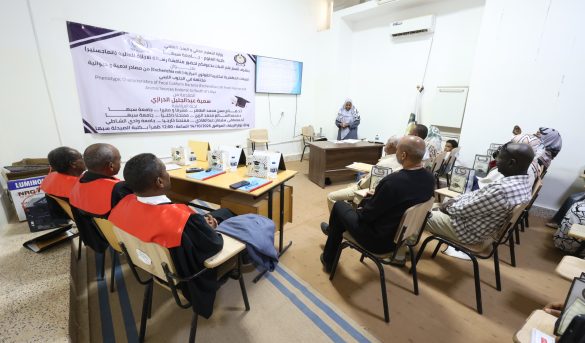 Audience seated in a classroom watching a presenter at a desk, with a large Arabic banner on the wall behind them.