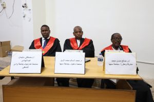Three judges in black robes with red sashes sit at a table with name placards in Arabic in a formal setting.