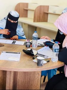 IMG-20261015-WA0008 - جامعة سبها | Sebha University Women wearing head coverings sit around a wooden round table, reviewing documents with pens, water bottles, and takeaway cups on the table.