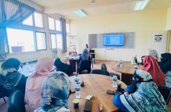 Women wearing hijabs seated around round tables in a classroom, facing a presenter and a large screen at the front.