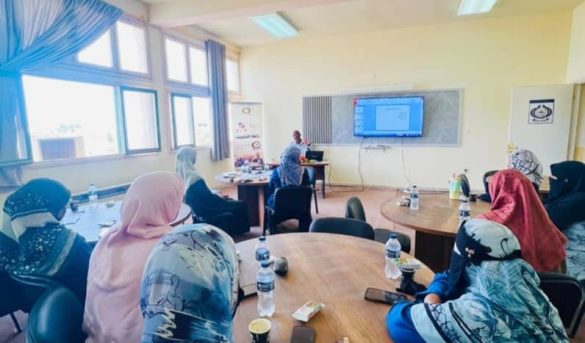 Women wearing hijabs seated around round tables in a classroom, facing a presenter and a large screen at the front.