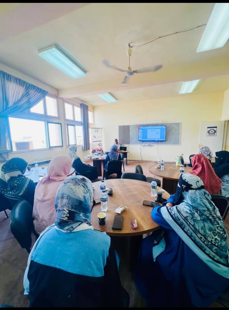 Women wearing hijabs seated around round tables in a classroom, facing a presenter and a large screen at the front.