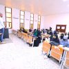 Male instructors stand at a podium at the front of a classroom while students in headscarves listen at desks.