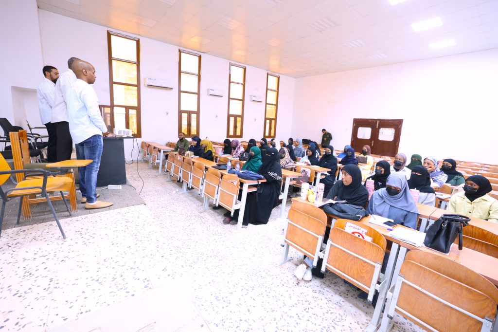 Male instructors stand at a podium at the front of a classroom while students in headscarves listen at desks.