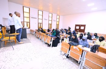 Male instructors stand at a podium at the front of a classroom while students in headscarves listen at desks.