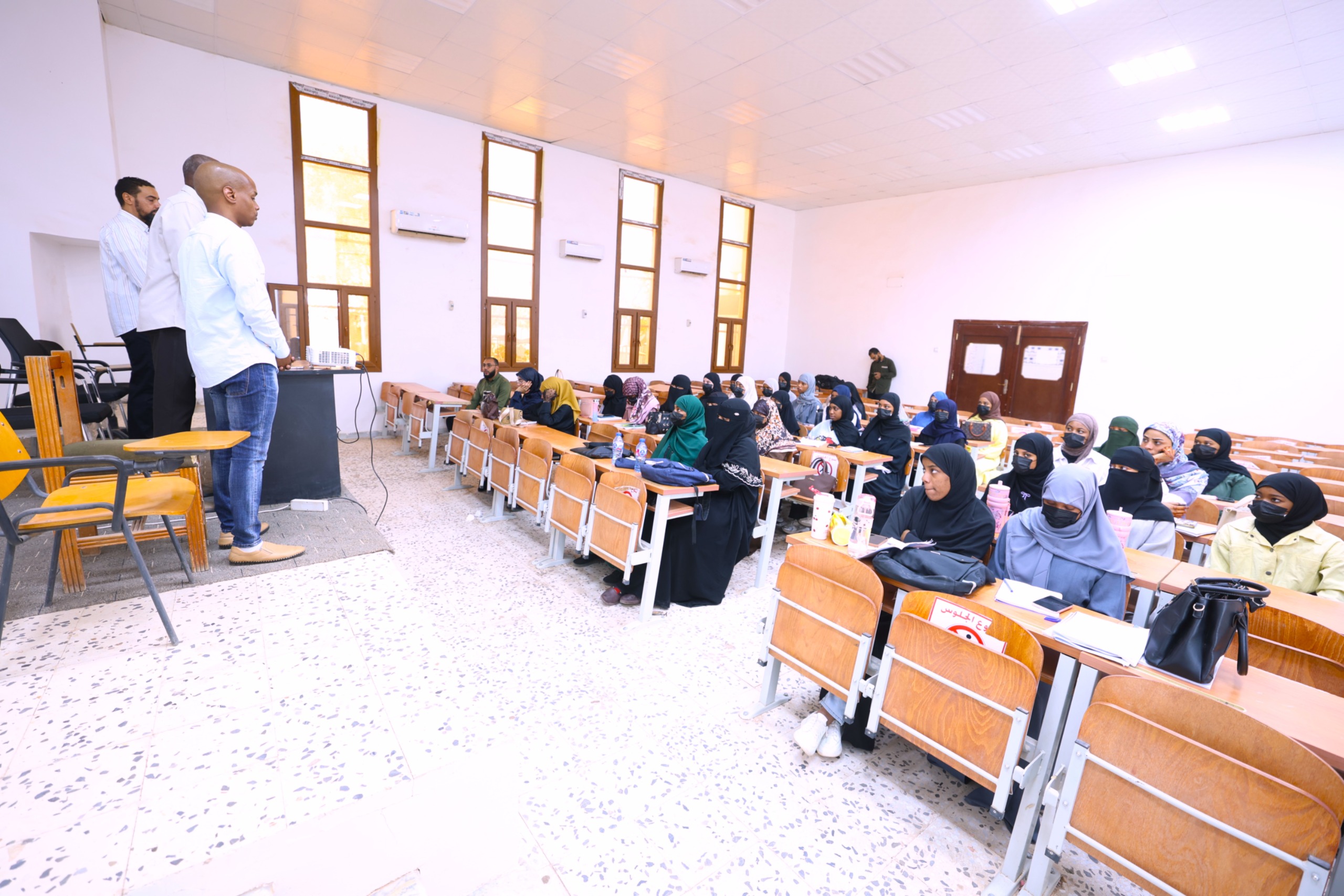 Male instructors stand at a podium at the front of a classroom while students in headscarves listen at desks.