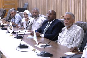 IMG-20261018-WA0025 - جامعة سبها | Sebha University Group of professionals seated at a long conference table with microphones in a meeting room, attentively listening.