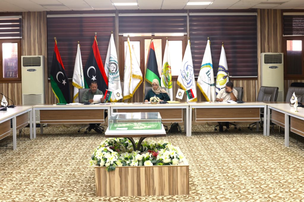 Conference room with multiple flagpoles and officials seated at a long table, center has a woman in hijab and a man reading documents, floral arrangement in foreground.
