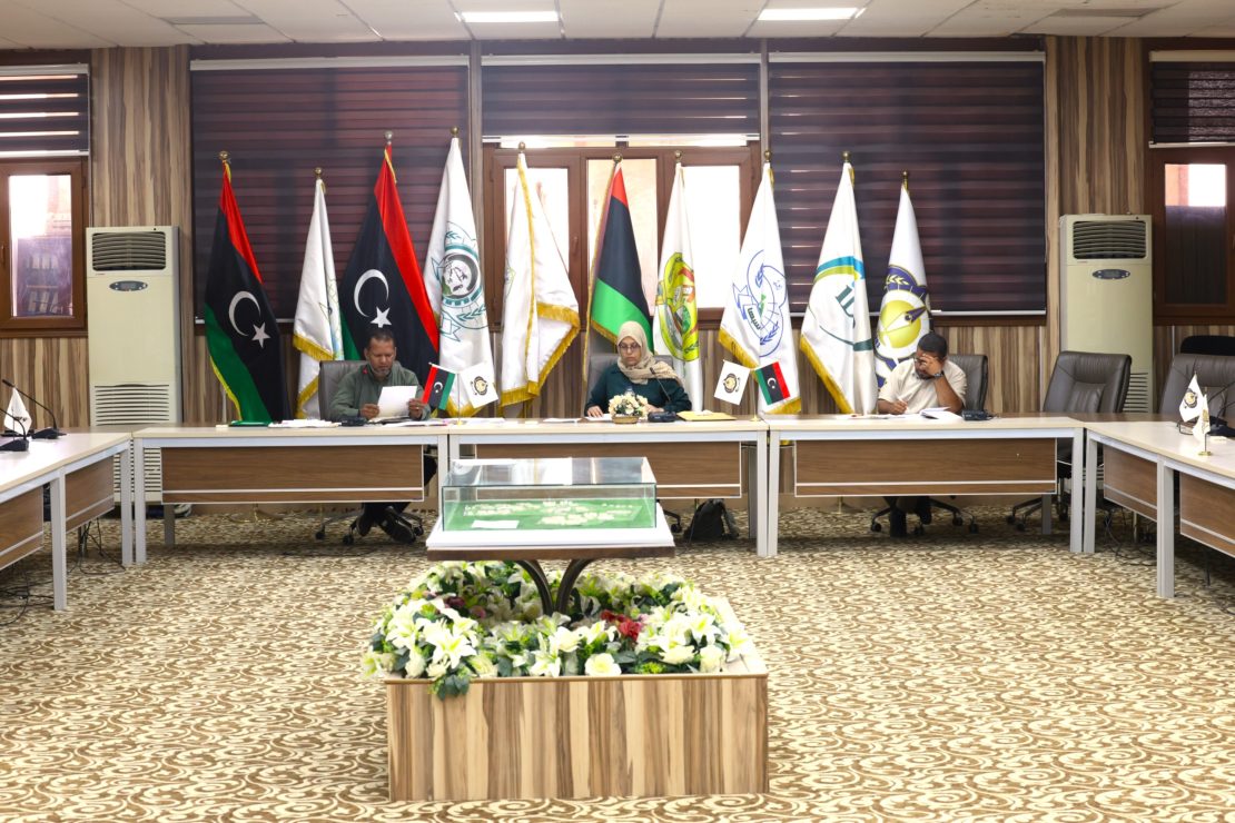 Conference room with multiple flagpoles and officials seated at a long table, center has a woman in hijab and a man reading documents, floral arrangement in foreground.