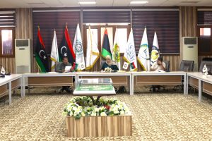 Conference room with multiple flagpoles and officials seated at a long table, center has a woman in hijab and a man reading documents, floral arrangement in foreground.