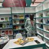 Man wearing a patterned shirt and red lanyard stands near a booth of green-and-white bookshelves filled with titles, with a table of yellow brochures and a smartphone in the foreground.