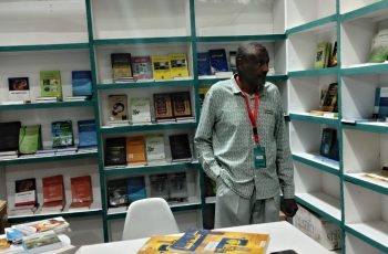 Man wearing a patterned shirt and red lanyard stands near a booth of green-and-white bookshelves filled with titles, with a table of yellow brochures and a smartphone in the foreground.