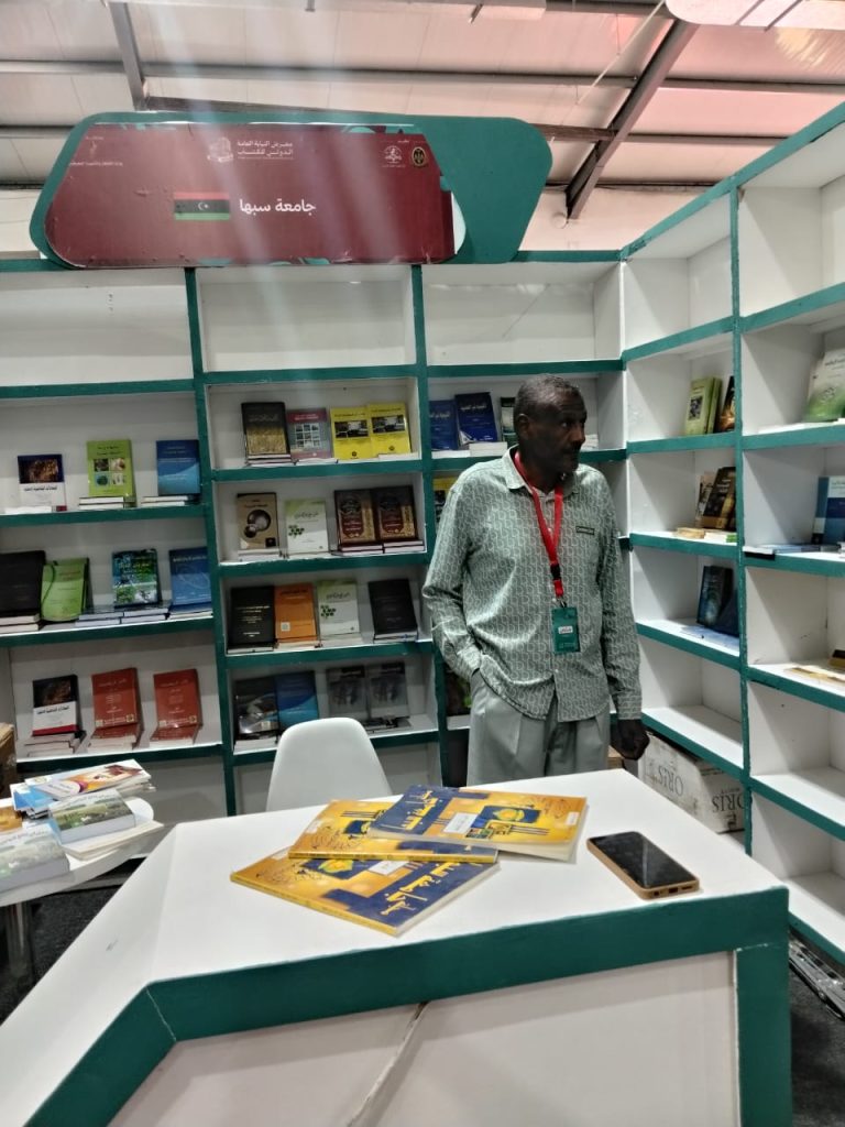 Man wearing a patterned shirt and red lanyard stands near a booth of green-and-white bookshelves filled with titles, with a table of yellow brochures and a smartphone in the foreground.