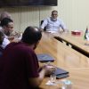 Group of men around a long wooden conference table during a meeting; flags displayed on the right and a bearded man at the head leads the discussion.