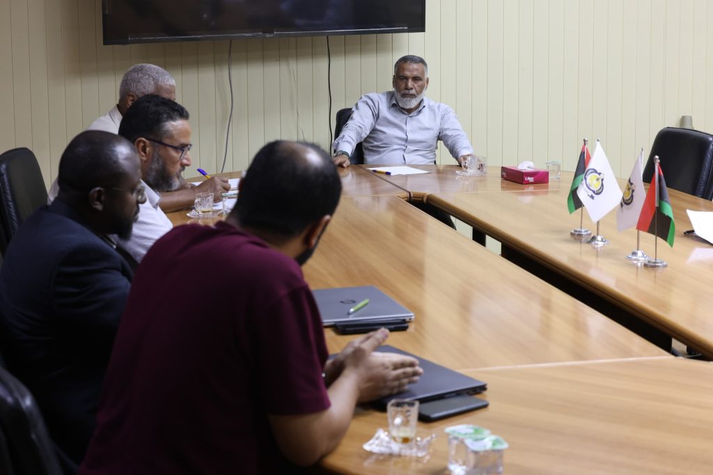 IMG-20261018-WA0077 - جامعة سبها | Sebha University Group of men around a long wooden conference table during a meeting; flags displayed on the right and a bearded man at the head leads the discussion.
