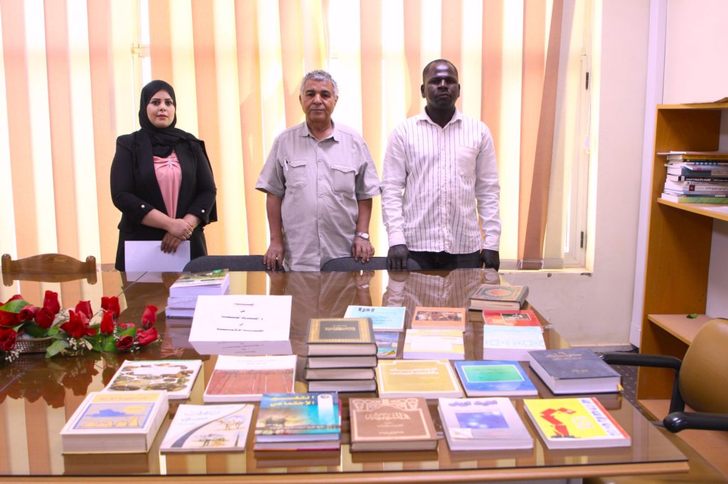 IMG-20261019-WA0010 - جامعة سبها | Sebha University Three adults stand behind a glass conference table covered with books, with red roses in the foreground.