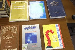 Row of Arabic books on a wooden shelf, featuring a yellow-covered title on the left, a blue book with a green square in the middle, and a dark blue hardcover on the right.