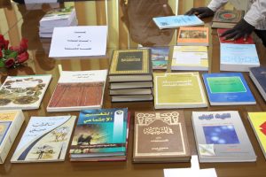 Table displaying numerous Arabic books on a glass surface, with colorful covers and a hand resting at the top-right corner.
