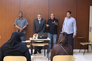 Four men stand at the front of a classroom giving a presentation to women wearing headscarves.