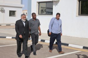 Three men walk together along a street beside a white building on a sunny day.