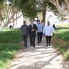 Group of men walking along a brick path under large trees in a sunny park setting.
