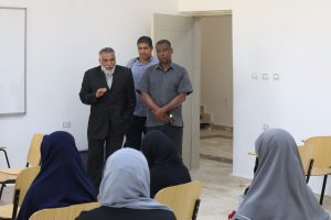 Three men stand at the front of a classroom addressing seated students wearing headscarves (hijab).
