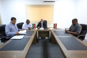 Four men sit around a conference table in a bright office, papers scattered, with Libyan flags on the table and a window with blinds behind them.