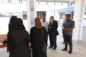 Group of five adults in a bright building corridor, two women in black abayas facing away, three men chatting near a pillar.