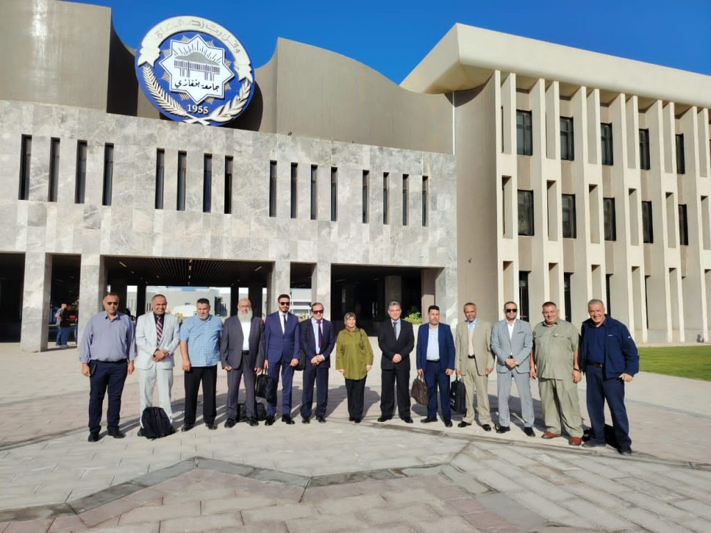 Group of professionals in business attire standing in a line outside a modern building with a circular emblem above the entrance (likely a university).