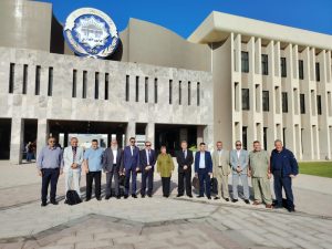 Group of professionals in business attire standing in a line outside a modern building with a circular emblem above the entrance (likely a university).