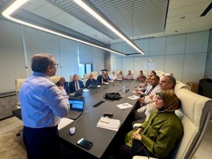 A presenter stands at the head of a long conference table addressing a diverse group of professionals seated around the table in a modern boardroom.