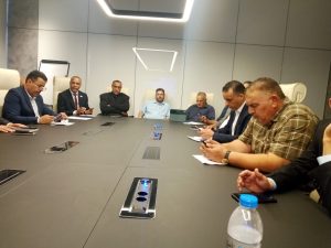 Group of men in a modern boardroom around a long conference table, some looking at phones during a meeting
