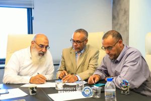 Three men sit at a conference table signing papers; left man with white beard, middle man in a tan suit, right man in a light purple shirt; university seal on table edge.