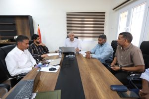 Six professionals sit around a wooden conference table in a boardroom, with laptops, papers, and a water bottle on the table amid a meeting.